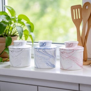May include: Three ceramic butter dishes with lids. Each dish is white with a swirled marble design in black, blue, and pink. The word "Butter" is embossed on the front of each dish. The dishes are on a white windowsill.