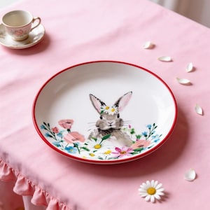 May include: White ceramic plate with a red rim, featuring a watercolour illustration of a grey bunny wearing a daisy crown, surrounded by colourful flowers. A teacup and saucer are in the background.