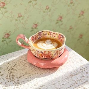 Puede incluir: Una taza de té con estampado floral con un asa rosa y un platillo rosa a juego. La taza está llena de un café con leche, cubierta con un diseño de espuma blanca. La taza y el platillo están sobre un mantel de encaje blanco.