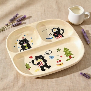 Pode incluir: Um prato bege dividido com ilustrações de gatos de desenho animado. O prato tem três seções e é decorado com imagens de gatos pretos, árvores e alimentos. Um pequeno jarro branco está ao lado do prato.