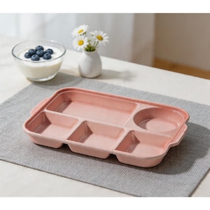 May include: A pink divided plate with several compartments for food. The plate is rectangular with rounded corners and a circular section. A small glass bowl of yogurt with blueberries and a vase of daisies are in the background.