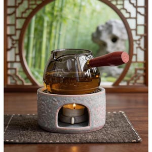 May include: A glass teapot with a wooden handle sits atop a stone warmer with a lit candle. The teapot contains tea and is set against a backdrop of a bamboo forest and a decorative window.
