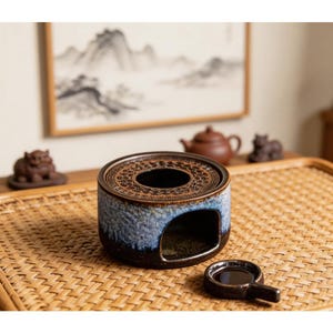 Puede incluir: Un calentador de té de cerámica con un esmalte azul y marrón, con un diseño circular en la parte superior y una abertura lateral. Una pequeña taza a juego está cerca. El calentador está sobre una bandeja tejida, con una tetera y una obra de arte en el fondo.