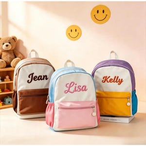 May include: Three backpacks with white front panels and personalized names embroidered in cursive. The left bag is brown, the center bag is pink and light blue, and the right bag is purple and yellow. Each features a front zippered pocket.