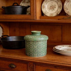 May include: A green ceramic butter dish with a lid, featuring a repeating floral pattern in a lighter shade of green. The dish is cylindrical and sits on a wooden surface, with other kitchenware visible in the background.