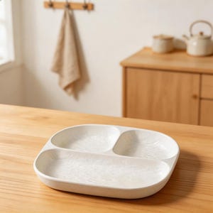 Könnte beinhalten: Eine weiße Servierplatte mit drei Fächern. Die Platte hat eine strukturierte Oberfläche und abgerundete Kanten. Sie steht auf einem Holztisch, mit neutralem Hintergrund.