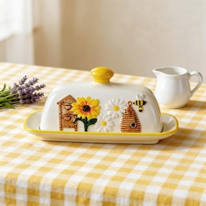 Może przedstawiać: Biała ceramiczna maselnica z żółtym guzikiem i wykończeniem, ozdobiona słonecznikiem, stokrotkami, ulem i pszczołą. Naczynie stoi na żółto-białej kraciastej serwetce. Po prawej stronie znajduje się mały biały dzbanek.