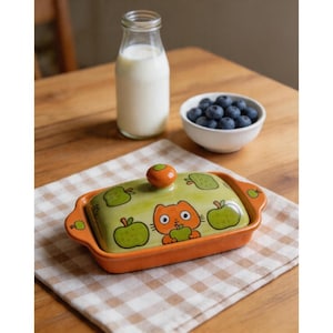 Könnte beinhalten: Rechteckige Butterdose aus Keramik in Orange und Grün mit Deckel, verziert mit einer Katze und Äpfeln. Auf einem karierten Tuch stehen eine Milchflasche und eine Schale mit Blaubeeren.