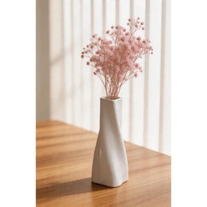 Pode incluir: Um vaso branco, moderno, com um design torcido, contém um buquê de flores delicadas, rosa claro. O vaso está sobre uma superfície de madeira, com luz natural iluminando a cena. O arranjo floral adiciona um toque de elegância.