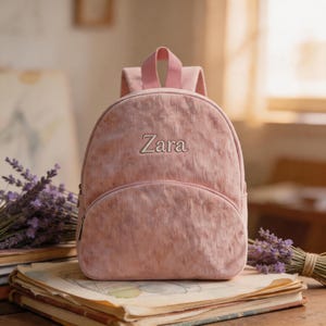 May include: A pink corduroy backpack with the name "Zara" embroidered on the front. The backpack has a small front pocket and a top handle. The background includes books and lavender.