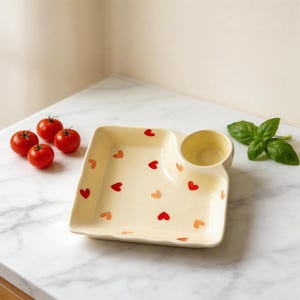 May include: A cream-coloured ceramic serving dish with a small, round dip bowl. The dish is decorated with red and peach-coloured heart shapes. Fresh tomatoes and basil leaves are arranged nearby, suggesting a food-related theme.