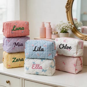 May include: A selection of cosmetic bags in a range of colours, such as red, purple, yellow, blue, and white. Each bag has a gingham or quilted pattern and is embroidered with a name in a contrasting colour. The bags are displayed on a white surface.