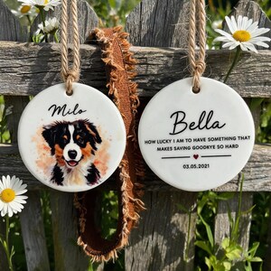 Op de afbeelding: Twee ronde witte keramische ornamenten hangend aan jute touw. Eén toont een aquarelillustratie van een zwart, wit en bruin hondje met de naam Milo. De andere toont de naam Bella, een herdenkingscitaat en de datum 03.05.2021.