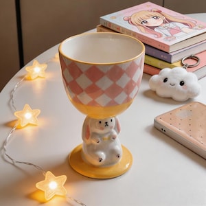 Puede incluir: Un vaso de cerámica caprichoso con un patrón de cuadros rosas y blancos. El vaso está sostenido por una figura de conejito blanco con orejas rosas y una base amarilla. Una cadena de luces en forma de estrella y libros están en el fondo.