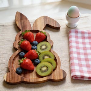 May include: A wooden bunny-shaped serving tray filled with fresh strawberries, blueberries, and kiwi slices. A white egg cup holds a pastel-colored egg. A pink and white checkered napkin is placed to the side.