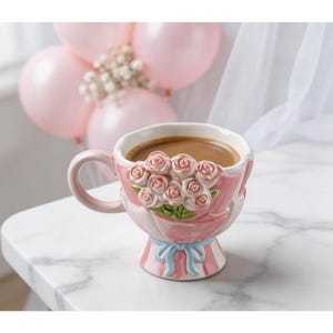 Może przedstawiać: Różowy ceramiczny kubek do kawy z motywem bukietu róż. Kubek ma uchwyt i podstawę z niebieskimi wstążkami. Kubek jest wypełniony brązowym płynem, prawdopodobnie kawą lub herbatą.