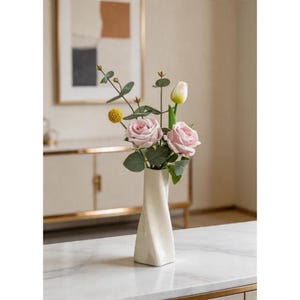 Pode incluir: Um vaso de cerâmica branco torcido com um arranjo floral. O arranjo inclui rosas cor-de-rosa, uma tulipa branca e detalhes amarelos e verdes. O vaso está sobre uma superfície com padrão de mármore.