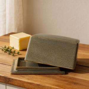 Peut inclure: Un beurrier rectangulaire en céramique de couleur vert-gris, avec le couvercle légèrement entrouvert. Un cube de beurre jaune et des brins d'herbes sont posés à côté du plat sur une surface en bois. Le plat a un design simple et moderne.