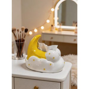 May include: A ceramic tissue box shaped like a crescent moon and cloud. The moon is yellow, and the cloud is white with yellow star accents. The tissue box is on a white surface, with makeup brushes in a glass and a vanity mirror in the background.