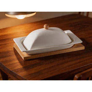 May include: A white ceramic butter dish with a wooden knob on top, resting on a rectangular wooden base. The dish has a rectangular shape with rounded edges. The butter dish is on a dark wooden table.