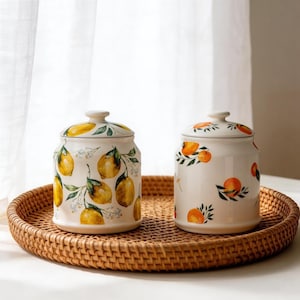 Op de afbeelding: Twee keramische potten met deksels, versierd met gele citroenen en groene bladeren, staan op een gevlochten dienblad. De potten hebben een witte achtergrond en worden waarschijnlijk in de keuken gebruikt.