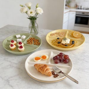 May include: Three divided plates in green, yellow, and white, each filled with different foods. The green plate holds mini cupcakes, marshmallows, and pretzels. The yellow plate has pasta, blueberries, and orange slices. The white plate contains a croissant, grapes, and boiled eggs.