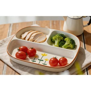 May include: A cream-coloured ceramic plate with a brown rim, divided into three sections. The plate contains sliced chicken, broccoli florets, and cherry tomatoes. Yellow flower illustrations decorate the plate.