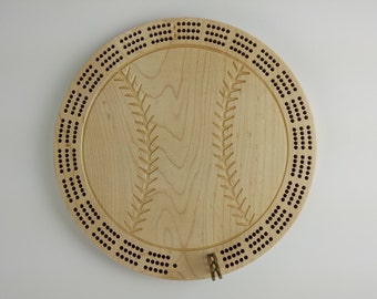 May include: A round wooden cribbage board designed to resemble a baseball. The board is light-colored with a baseball stitch design carved into the wood. It has numerous holes around the perimeter for pegging and includes metal pegs.