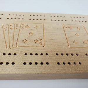 May include: A wooden cribbage board with a light brown finish. The board has two sets of cards engraved on it, one set of four cards with a Jack and three fives, and one card with a five of diamonds. The board has a row of holes along the bottom.