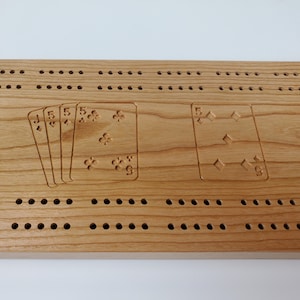 May include: A wooden cribbage board with a natural wood finish. The board has a row of holes for pegs and a design of playing cards, including a Jack, three fives, and a five of diamonds.