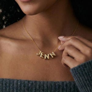 以下が含まれることがあります： ブロック体の「JENN」という名前が入ったゴールドカラーのネックレス。ネックレスは首にかけられています。その人はグレーのセーターを着ています。