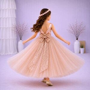 May include: A floor-length rose gold dress with a V-neckline and a large sequined bow at the waist. The skirt is made of layered tulle, and the bodice features matching sequins. A floral headband with small pink flowers is worn on the hair.