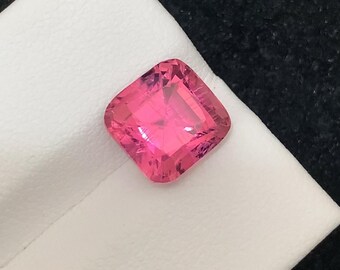 May include: A vibrant, cushion-cut pink gemstone rests on a white surface against a black background. The gemstone's facets reflect light, showcasing its brilliant color. This gemstone is a beautiful example of fine jewelry.