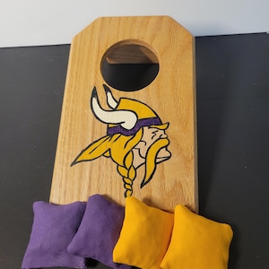 May include: Wooden cornhole board featuring the Minnesota Vikings logo in yellow, purple, and black. Includes four bean bags, two purple and two gold. The board has a hole for tossing the bean bags.