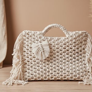 Può includere: Borsa a mano in macramè color crema con forma rettangolare e motivo intrecciato. La borsa presenta un manico, frange e un accento decorativo a forma di piuma. La borsa è realizzata in una fibra naturale e morbida.