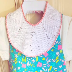 Könnte beinhalten: Ein weißes Strick-Lätzchen mit rosa Besatz, über einem Kleid mit Blumenmuster mit einem weißen Oberteil mit kurzen Ärmeln. Das Kleid hat einen blauen Hintergrund mit rosa und gelben Blumenmustern. Das Outfit hängt an einem weißen Kleiderbügel.