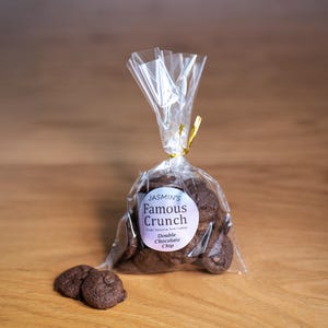 May include: A transparent cellophane bag tied with a gold ribbon, filled with chocolate chip cookies. The bag has a circular label that reads "Jasmin's Famous Crunch" and "Double Chocolate Chip". Two cookies are in front of the bag.