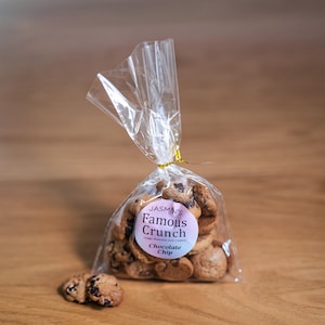 May include: A cellophane bag tied with gold ribbon, filled with chocolate chip cookies. The bag has a round label that reads "Jasmin's Famous Crunch Chocolate Chip". One cookie rests in front of the bag on a wooden surface.