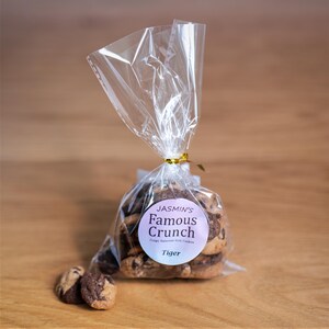 May include: A transparent cellophane bag, tied with a gold ribbon, holds several chocolate chip cookies. The bag has a round sticker that reads "Jasmin's Famous Crunch" and "Tiger". Two cookies are outside the bag on a wooden surface.