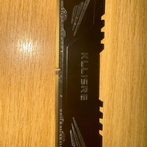 May include: A black KLLISRE RAM stick with gold-colored connectors. The RAM stick has a textured surface with diagonal lines and the brand name printed in white. The RAM stick is on a wooden surface.
