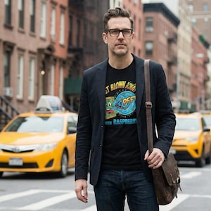 Könnte beinhalten: Ein Mann trägt ein schwarzes T-Shirt mit einem farbenfrohen Grafikdesign und dem Text "GET BLOWN AWAY WITH RASPBERRY". Er trägt einen marineblauen Blazer, blaue Jeans und eine Brille und hat eine braune Umhängetasche dabei. Gelbe Taxis sind im Hintergrund.