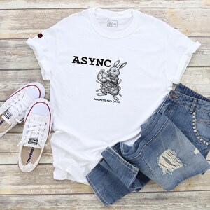 Peut inclure: T-shirt blanc avec le mot "ASYNC" et un dessin de lapin tenant une horloge, associé à un jean bleu et des baskets blanches. Le texte "AWAITS NO ONE" est sous le dessin du lapin. L'ensemble est posé sur une surface en bois.