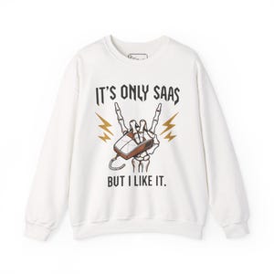 Peut inclure: Sweat-shirt blanc avec un motif représentant une main de squelette tenant une souris d'ordinateur en signe de rock and roll. Le texte dit "IT'S ONLY SAAS BUT I LIKE IT" en noir, avec des éclairs dorés.