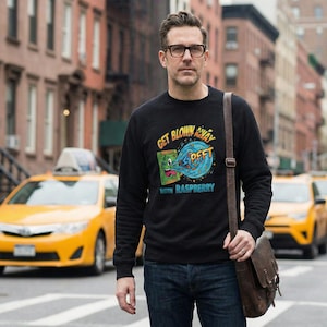 Op de afbeelding: Zwart sweatshirt met de tekst "GET BLOWN AWAY" en "PFFT" met een afbeelding van een boek en golven. Het sweatshirt heeft ook de tekst "WITH RASPBERRY". De persoon draagt een bril en een bruine leren schoudertas.