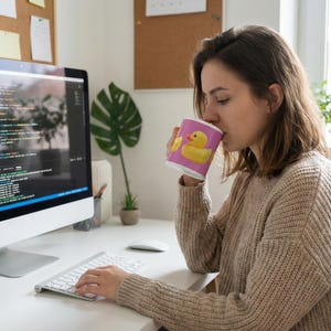 May include: A white coffee mug featuring a pink background and yellow rubber duck design. The mug is held by a person at a desk with a computer and keyboard. The person is wearing a beige sweater.