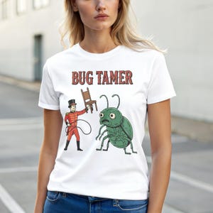 Può includere: T-shirt bianca con la scritta "BUG TAMER" in rosso. La grafica mostra un personaggio dei cartoni animati in uniforme rossa che tiene una frusta e una sedia, di fronte a un grosso insetto verde con un'espressione preoccupata.