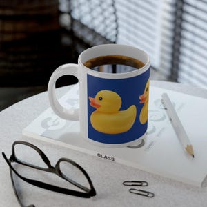 May include: White ceramic mug with a yellow rubber duck design on a blue panel. The mug is filled with coffee and sits on a white surface with a pencil, glasses, and paperclips.