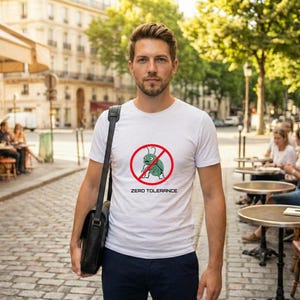 Puede incluir: Una camiseta blanca con un gráfico de un insecto verde tachado dentro de un círculo rojo, con las palabras "ZERO TOLERANCE" debajo. La persona lleva un bolso negro y pantalones azul oscuro.