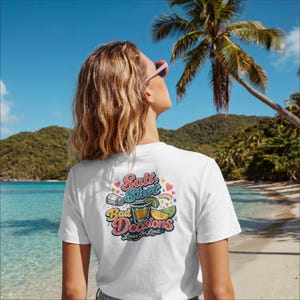 Puede incluir: Camiseta blanca con un gráfico colorido en la espalda que dice "Salt Shot Bad Decisions Limes The Limit". El gráfico incluye un salero, un vaso de chupito, una lima y un arcoíris. La camiseta se lleva en una playa.