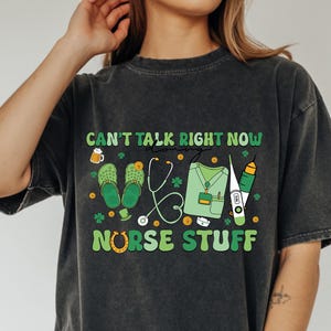 May include: Dark gray t-shirt with the text "CAN'T TALK RIGHT NOW NURSE STUFF" in green and yellow. The graphic includes medical tools, clogs, and shamrocks. The shirt is a casual, oversized fit.